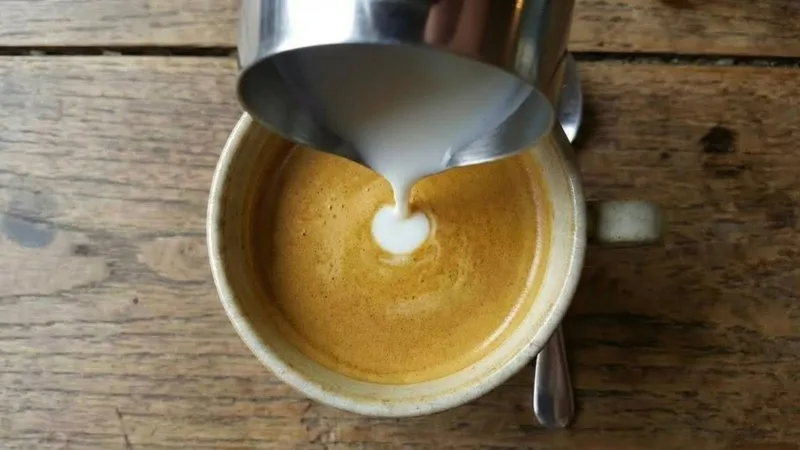 Lowering the pitcher tip to nearly touch the surface, causing the white milk to stay on top of the crema and form the initial Monk's Head circle